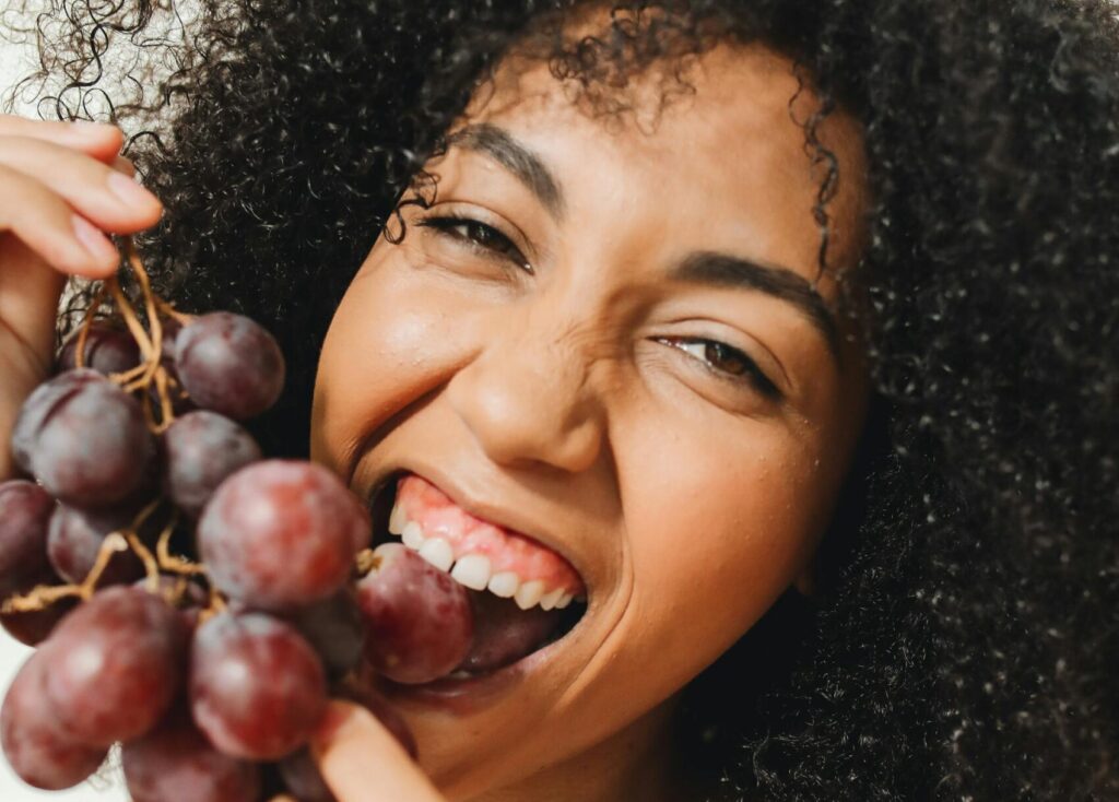 Portrait of a smiling woman eating red grapes indoors, conveying joy and freshness.