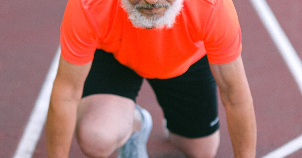 Diligent senior male looking at camera and standing in crouch start on track on blurred background of stadium
