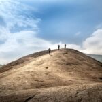 Three hikers trekking up a large rocky hill under a clear blue sky, surrounded by lush greenery.
