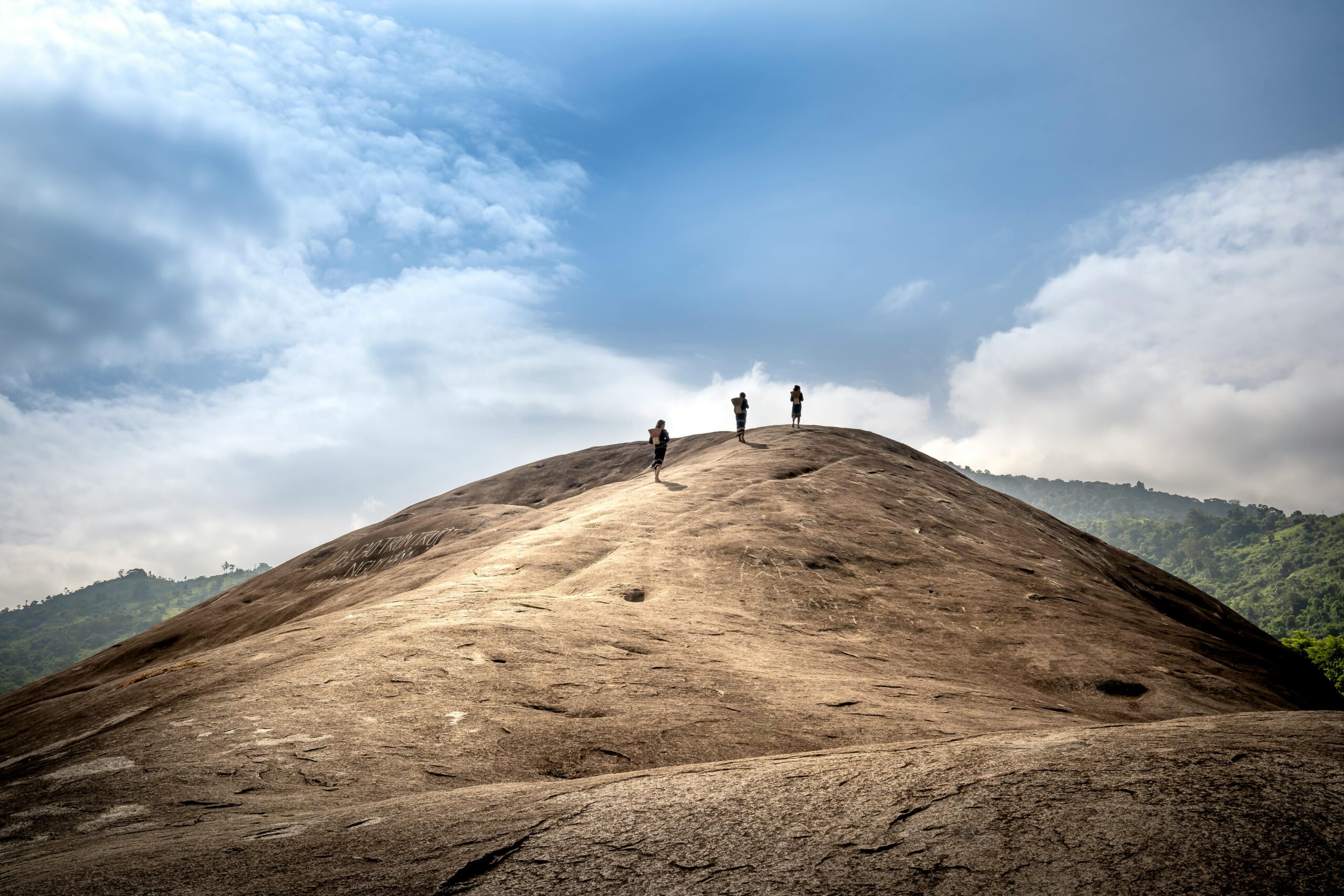 Three hikers trekking up a large rocky hill under a clear blue sky, surrounded by lush greenery.